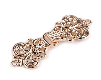 May include: A gold-colored metal clasp with an ornate, swirling design. The clasp features a hook and eye closure and is likely used for jewelry or garment embellishment. The intricate details and polished finish suggest a vintage or antique style.