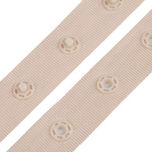 Puede incluir: Dos correas de tela beige con cierres de presi&oacute;n blanquecinos espaciados uniformemente. Las correas est&aacute;n en &aacute;ngulo sobre un fondo blanco, mostrando la textura del material tejido y los detalles de los cierres de presi&oacute;n.