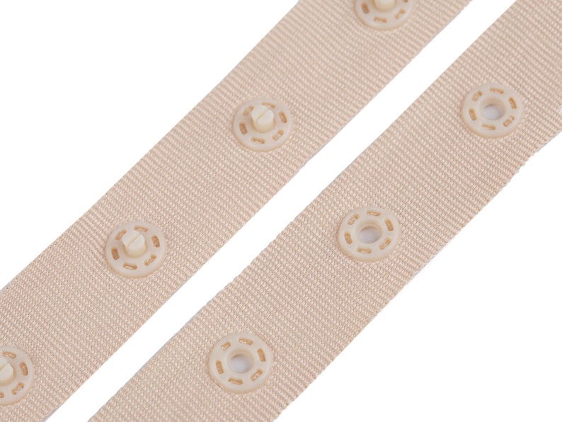 Puede incluir: Dos correas de tela beige con cierres de presi&oacute;n blanquecinos espaciados uniformemente. Las correas est&aacute;n en &aacute;ngulo sobre un fondo blanco, mostrando la textura del material tejido y los detalles de los cierres de presi&oacute;n.