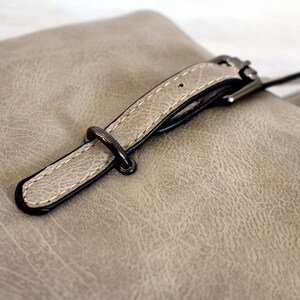 May include: A close-up of a light brown leather handbag with a black buckle and strap.