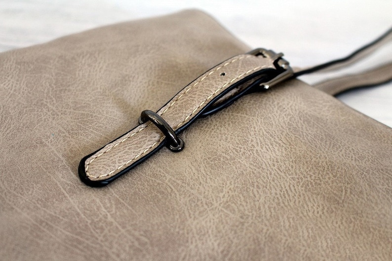 May include: A close-up of a light brown leather handbag with a black buckle and strap.