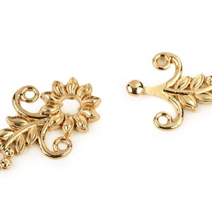 Op de afbeelding: Twee gouden metalen decoratieve versieringen met een bloemmotief. Elke versiering heeft een gestileerde bloem met bladeren en een lus om te bevestigen.