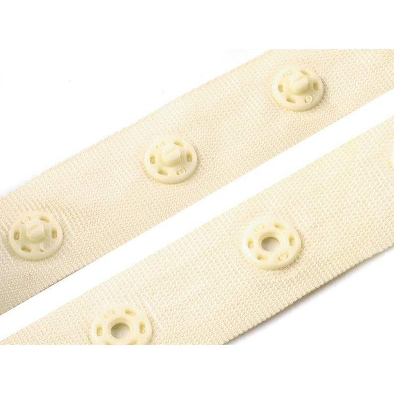 Puede incluir: Primer plano de dos tiras de tela blanquecina con broches de presi&oacute;n color crema espaciados uniformemente. Los broches tienen un dise&ntilde;o circular con un tornillo central. La tela tiene una textura tejida.