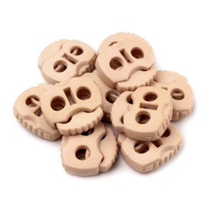 Peut inclure: Un tas de boutons décoratifs beiges. Chaque bouton a une forme unique avec un trou central et un bord festonné. Les boutons sont regroupés sur un fond blanc.