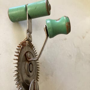 以下が含まれることがあります： 酸化の兆候が見られる金属製のギア機構を備えた、ヴィンテージの手回し式回転泡立て器。このキッチンツールには、上部とサイドのクランクアームにそれぞれ1つずつ、ライトグリーンに塗装された木製のハンドルが付いています。