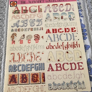 May include: A cross-stitch pattern booklet titled The Alphabet Collection Vol. 3 BK-42. The cover displays various embroidered alphabet styles in multiple colors, including floral, animal, and geometric motifs, arranged in rows on a cream background.