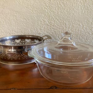 May include: A clear glass casserole dish with a lid and a silver metal serving dish. The casserole dish has a rounded shape and a clear glass lid with a small handle. The silver dish has handles and a decorative openwork design.
