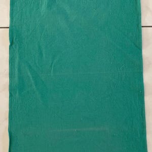 Può includere: Un panno rettangolare verde smeraldo con una superficie strutturata. Il tessuto sembra essere piegato e ha un aspetto leggermente stropicciato. Il panno è probabilmente un asciugamano da cucina o da bagno.