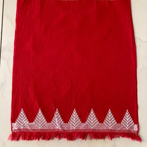 May include: A rectangular red textile featuring white embroidered geometric patterns along the top and bottom edges. The design includes a series of repeating triangular motifs at the base, finished with a fringed edge at both the top and bottom.
