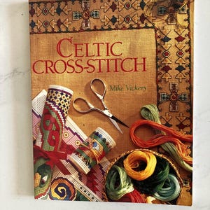 May include: A book titled Celtic Cross-Stitch by Mike Vickery featuring a cover with a brown textured background, a pair of silver embroidery scissors, two rolled cross-stitch patterns, and a small basket filled with colorful embroidery floss.
