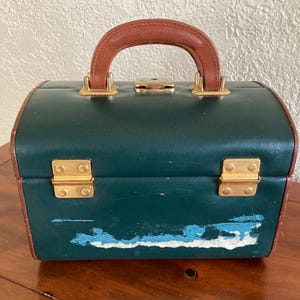 May include: A vintage green vanity case with a brown leather handle and trim. The case has gold-colored metal clasps and a latch. The paint on the front of the case is chipped, revealing a blue undercoat. The case is sitting on a wooden surface.