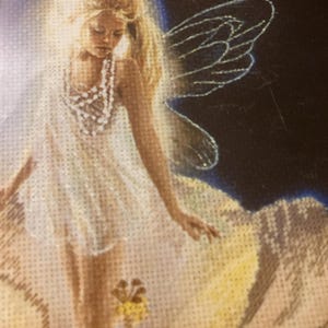 May include: A cross-stitch embroidery depicting a fairy with long blonde hair, wearing a white dress and a beaded necklace. The figure has translucent wings and is surrounded by a glowing light against a dark background, with small yellow flowers.