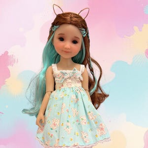 Puede incluir: Una muñeca con cabello largo castaño y verde azulado que viste un vestido azul claro con un patrón repetitivo de pequeños animales blancos y flores. El vestido tiene un corpiño con volantes y un lazo, y un dobladillo de encaje blanco. Botas beige.