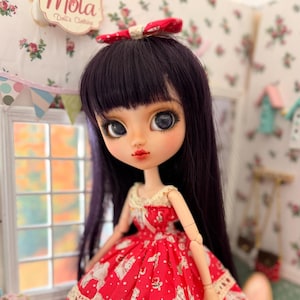 May include: A doll with long dark purple hair and large grey eyes wearing a red dress with a white polka dot and floral pattern. The dress features a cream-colored lace trim at the waist and hem, with a matching red bow in the doll's hair.