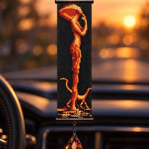 May include: A rectangular beaded car hanging ornament featuring a black background with a vertical orange and yellow abstract figure and a small animal silhouette. The piece is suspended by a black chain and features a faceted amber crystal drop.