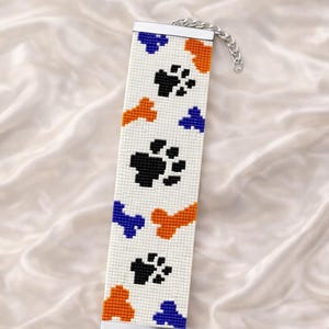 May include: A white beaded bracelet pattern with black paw prints, orange and blue shapes. The bracelet has a silver clasp and chain. Text at the top says "Bracelet Bead Pattern" and at the bottom "Loom Stitch".