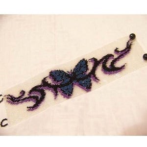 K&ouml;nnte beinhalten: Ein breites Perlenarmband mit wei&szlig;em Hintergrund und einem zentralen blauen Schmetterlingsmotiv. Das Design umfasst schwarze und violette, tribalartige Flammenmuster, die vom Schmetterling ausgehen. An den Enden sind zwei schwarze Perlen befestigt.