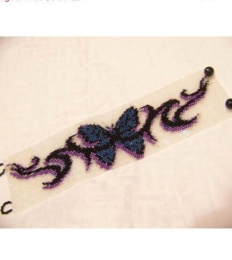 K&ouml;nnte beinhalten: Ein breites Perlenarmband mit wei&szlig;em Hintergrund und einem zentralen blauen Schmetterlingsmotiv. Das Design umfasst schwarze und violette, tribalartige Flammenmuster, die vom Schmetterling ausgehen. An den Enden sind zwei schwarze Perlen befestigt.