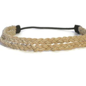 May include: A braided headband in a light tan color. The headband features two parallel rows of braided material, connected by a black elastic band. The braided design gives the headband a textured appearance.