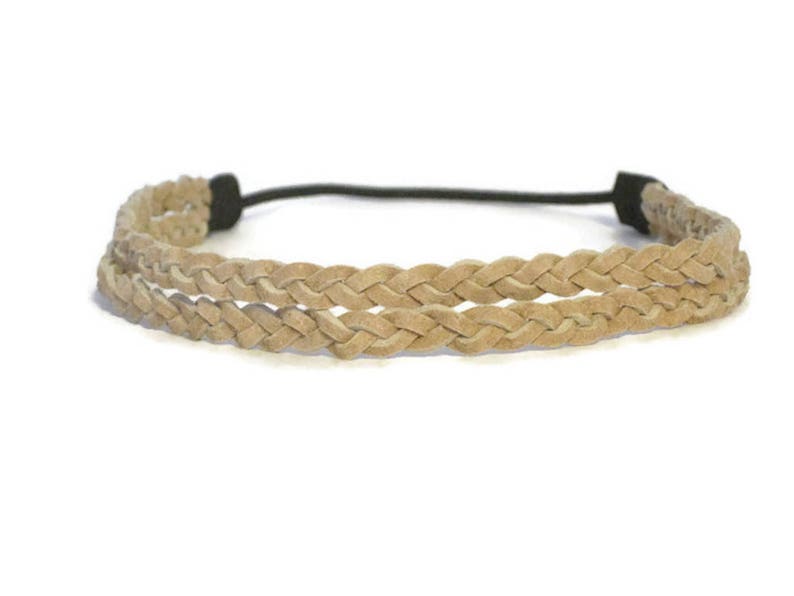 May include: A braided headband in a light tan color. The headband features two parallel rows of braided material, connected by a black elastic band. The braided design gives the headband a textured appearance.