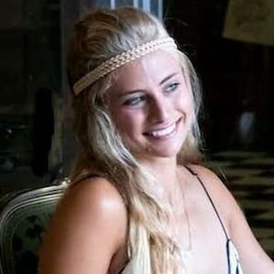 May include: A woman with long blonde hair and a braided headband smiles. She wears a spaghetti strap top. The background is blurred, suggesting an indoor setting. The image is well-lit, highlighting her features.