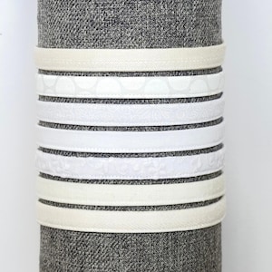 May include: A collection of eight thin headbands in various shades of white and cream, featuring different textures and patterns including solid, polka dot, floral, and paisley designs, stacked vertically on a gray fabric display cylinder.