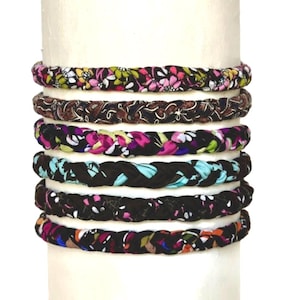 May include: A stack of six braided fabric headbands featuring various floral and abstract patterns in black, pink, green, blue, and orange. Each headband is constructed from a soft, woven material and is displayed on a cylindrical white form.
