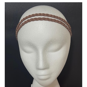 May include: A brown braided headband with two strands is displayed on a white mannequin head. The headband is a warm, neutral brown color and appears to be made of a soft material. The background is a dark gray.