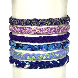 May include: A stack of seven braided fabric headbands in various colors and patterns. The headbands include floral and solid designs in shades of purple, blue, and green. The headbands are displayed on a white cylindrical form.