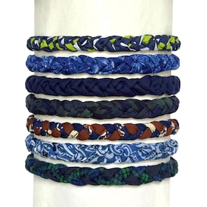 May include: A set of seven braided fabric bracelets stacked vertically. The bracelets feature various patterns and colors, including lime green and navy, solid navy, dark plaid, brown and navy floral, light blue paisley, and green plaid.