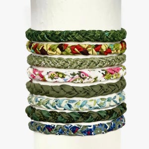 May include: A stack of nine braided fabric headbands featuring various patterns and colors, including shades of forest green, olive, floral prints with pink and white, and blue botanical designs, all arranged vertically on a cylindrical display.