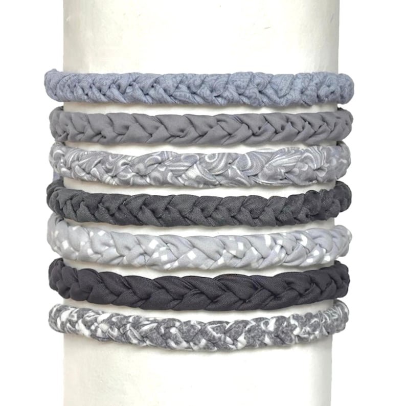 May include: A set of seven braided fabric headbands in various shades of gray. The collection includes solid gray, charcoal, and light gray tones, along with patterned designs featuring swirls, checks, and mottled textures in a soft textile material.