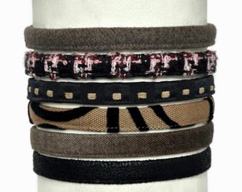 May include: A stack of six headbands featuring various textures and patterns, including a solid brown felt, a black tweed with pink flecks, a black band with tan squares, a tan band with black stripes, a solid brown velvet, and a black textured band.