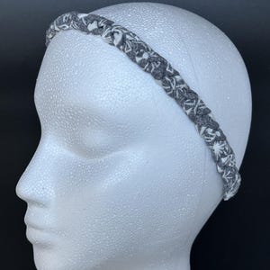 May include: A braided fabric headband featuring a dark gray and white patterned design. The headband is constructed from multiple strands of material woven together to create a textured, continuous loop that sits across the top of the head.