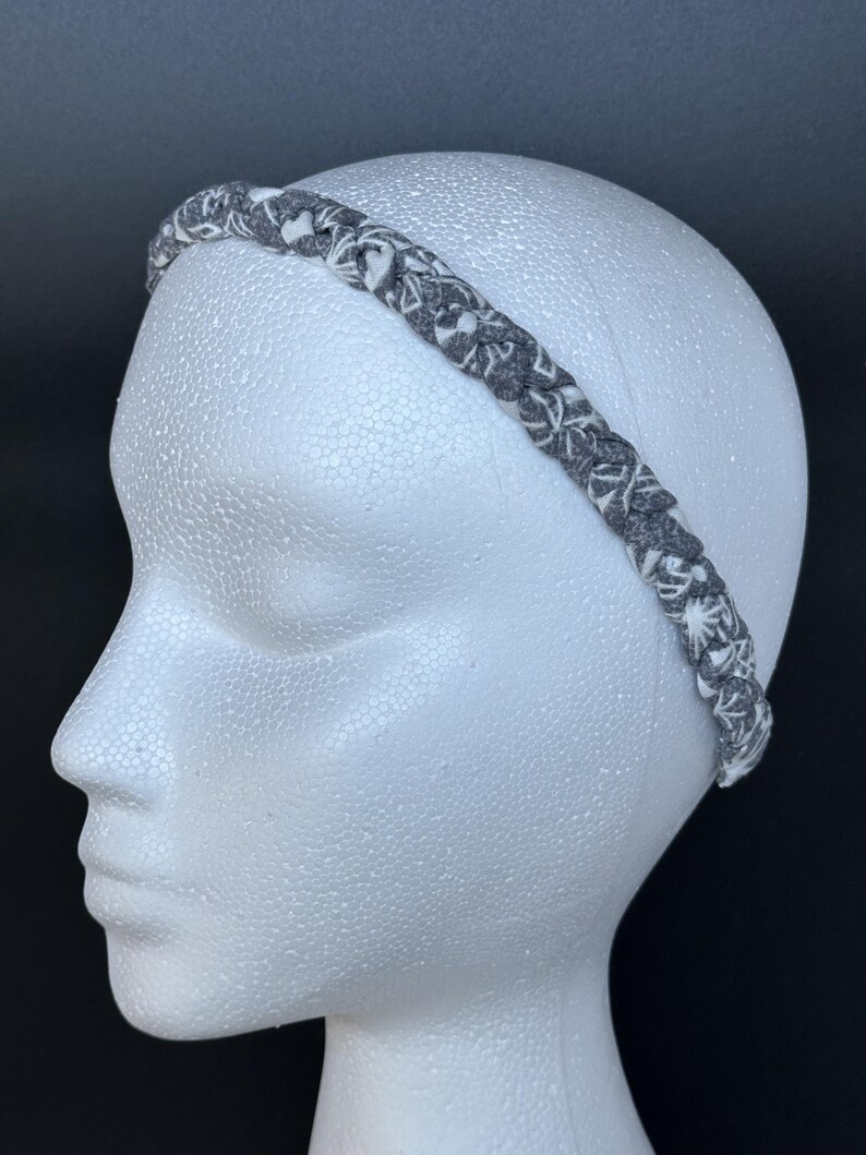 May include: A braided fabric headband featuring a dark gray and white patterned design. The headband is constructed from multiple strands of material woven together to create a textured, continuous loop that sits across the top of the head.