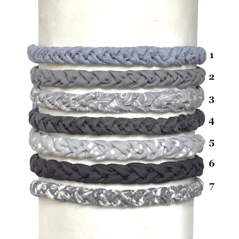 May include: Seven braided fabric headbands arranged vertically, numbered one through seven. The headbands feature various shades of gray, including solid colors, a light gray swirl pattern, a light gray checkered print, and a mottled gray texture.