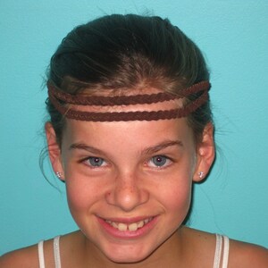 May include: A brown braided headband with three strands. The headband is worn across the forehead. The background is a solid turquoise color.