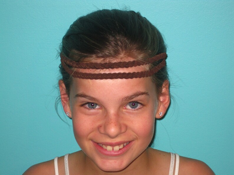 May include: A brown braided headband with three strands. The headband is worn across the forehead. The background is a solid turquoise color.