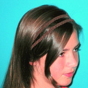May include: A brown braided headband worn in the hair. The headband is made of a thin, woven material and sits across the forehead. The hair is long, dark brown, and styled with a side part. The background is a solid turquoise color.