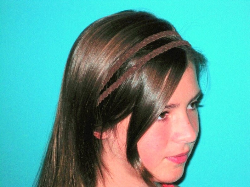 May include: A brown braided headband worn in the hair. The headband is made of a thin, woven material and sits across the forehead. The hair is long, dark brown, and styled with a side part. The background is a solid turquoise color.
