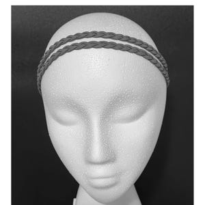May include: A gray braided headband, featuring two parallel bands, is displayed on a white foam mannequin head. The headband's texture and design are clearly visible against the neutral background, showcasing a simple yet stylish hair accessory.
