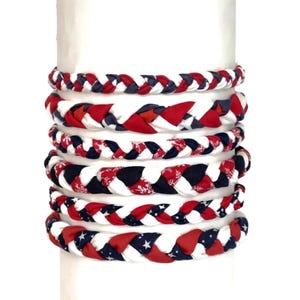 May include: Braided fabric bracelets in red, white, and blue. The bracelets feature patriotic patterns, including stars, snowflakes, and solid colors. The bracelets are stacked on a white cylindrical form.