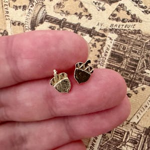 Peut inclure: Une paire de petites boucles d'oreilles à clous en forme de gland doré. Chaque boucle présente un capuchon texturé avec un motif hachuré et une base lisse et polie. Le design du gland est réalisé dans une finition dorée métallique.