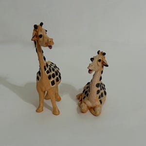 May include: Two giraffe figurines with light tan bodies and dark brown, blocky spot patterns. One giraffe is standing upright with its head turned slightly, while the other giraffe is in a seated position with its legs tucked underneath its body.