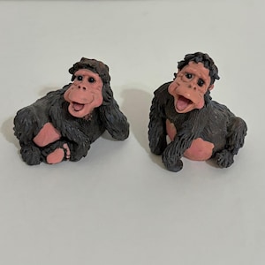 May include: Two small figurines of primates with dark brown textured fur and light pink skin on their faces, hands, and feet. One primate is in a reclining position, while the other is sitting upright with its mouth open in a wide expression.