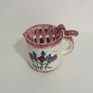 May include: A cream-colored ceramic flower frog vessel featuring a hand-painted floral motif with red roses and blue lavender sprigs. The top includes a removable grid lid with a side handle, all finished with a speckled maroon rim.