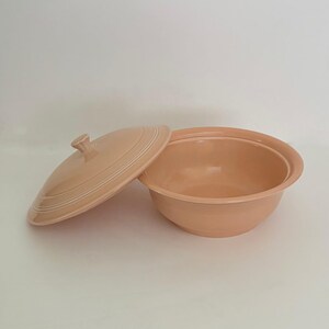 May include: A light peach-colored ceramic serving bowl with a matching lid. The bowl features a wide, circular opening and a series of concentric ring details around the exterior base. The lid has a central knob handle and matching ring details.