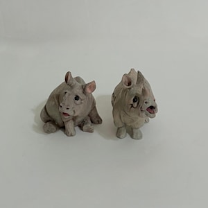 May include: Two small, grey rhinoceros figurines crafted with detailed textures. One rhinoceros is in a seated position, while the other stands with its mouth slightly open. Both figures feature dark eyes, pointed ears, and a single horn on the snout.
