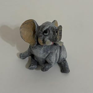 May include: A small, gray elephant figurine with a textured, stone-like finish. The elephant is depicted in a seated position with large, tan-colored ears, a raised trunk, and small white tusks. The facial features include dark, expressive eyes.