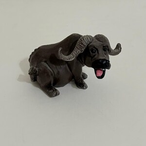 May include: A small, dark brown figurine of a water buffalo in a seated position. The animal features prominent curved horns, a black muzzle, and an open mouth revealing a pink tongue. The surface texture is matte with subtle molded details.
